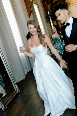 Smiling bride in white strapless gown holding a drink, walking with groom in black tuxedo at wedding reception.