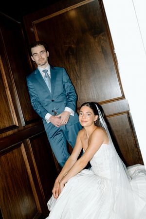 Bride in white gown and groom in blue suit pose elegantly in a wood-paneled doorway on their wedding day.
