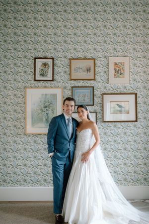 A bride and groom pose elegantly in front of floral wallpaper adorned with framed artwork on their wedding day.