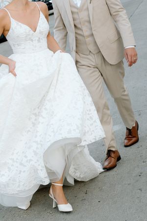 Close-up view of wedding party feet showing white bridal gowns and tan suits with brown leather dress shoes on pavement.