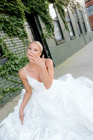 A person in a white wedding dress poses against an ivy-covered brick wall on a city street.