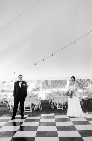 A wedding couple standing apart on a black and white checkered dance floor under a white tent.