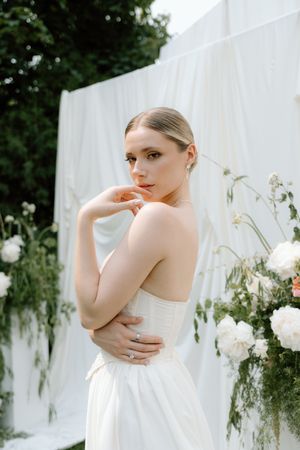 Elegant bride in strapless white gown poses outdoors with white floral arrangements and draped fabric backdrop.