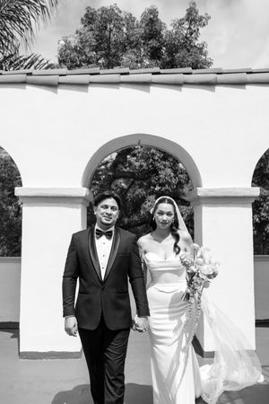 Black and white photo of a bride and groom holding hands, walking in front of a white arched wall with trees.
