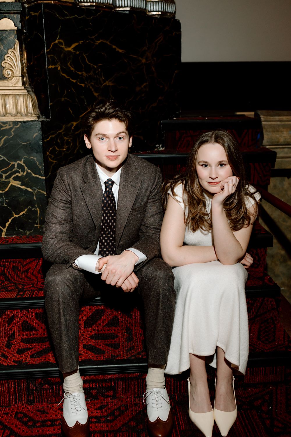 A young man in a gray suit and a young woman in a white dress sit together on red carpeted steps.