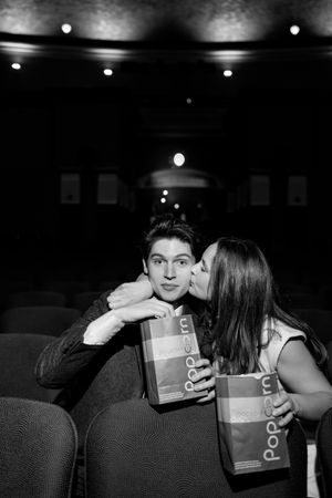 Couple sharing a romantic kiss in a dark movie theater, holding popcorn bags during a date night.