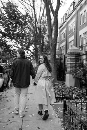 Black and white photo of a couple walking on a tree-lined sidewalk past elegant townhouses in autumn.