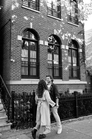 A romantic couple embraces outside a classic brick building with arched windows on a city street.