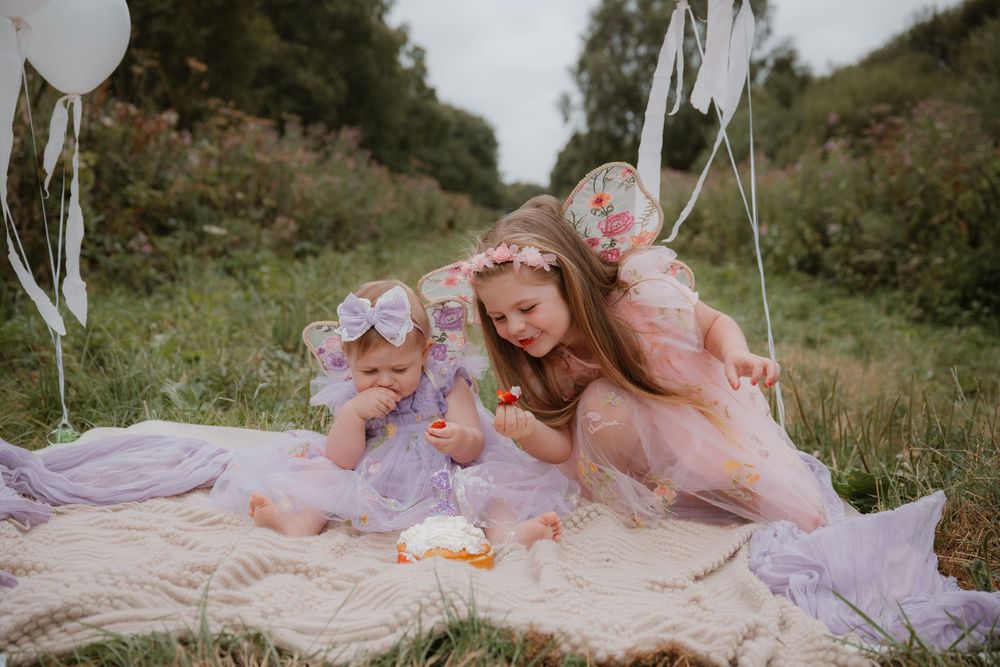 Three young girls in pastel fairy costumes sit on a blanket outdoors, sharing a sweet moment together.
