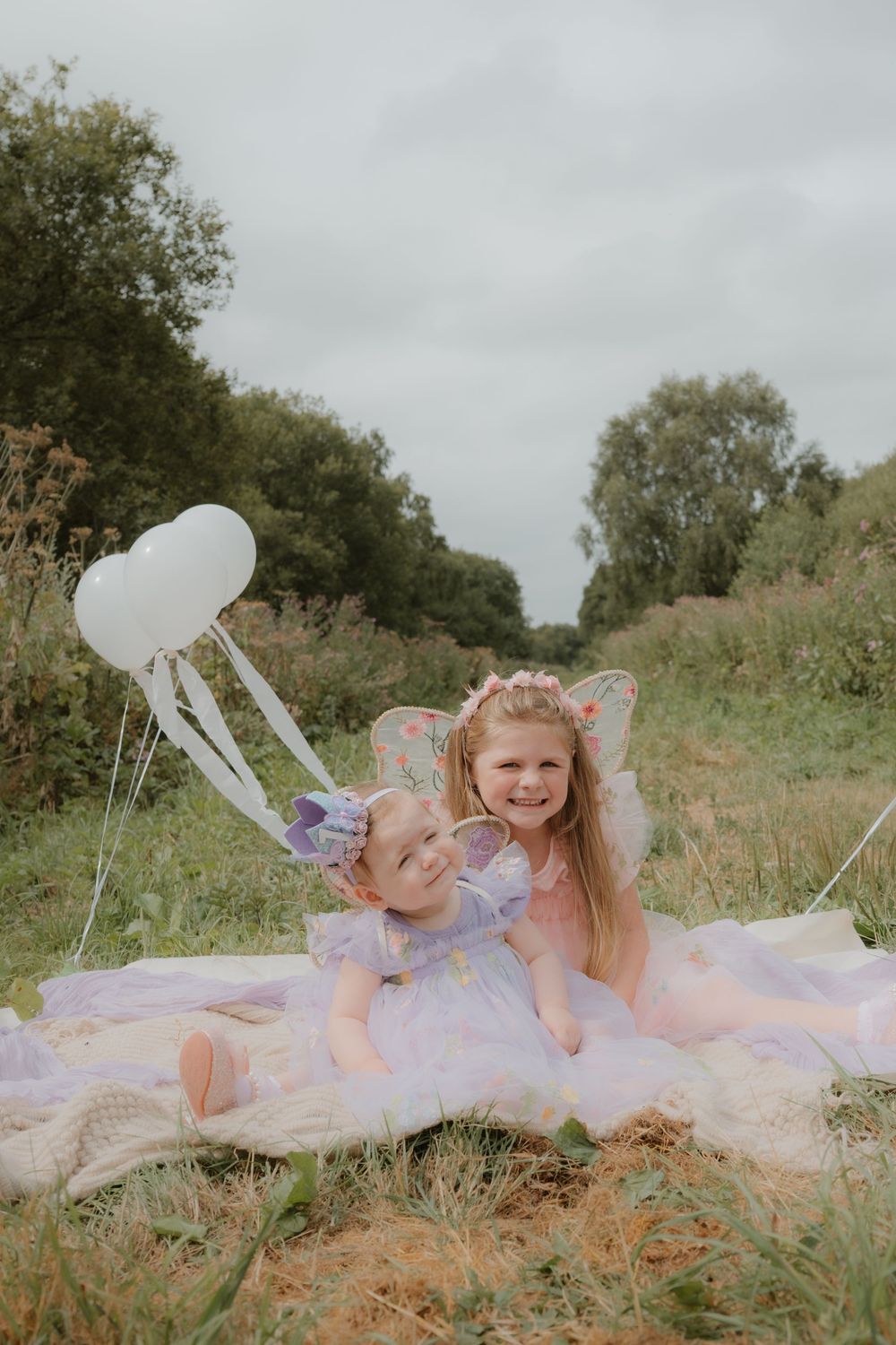 Two girls in fairy costumes with wings sit on a blanket in a meadow, holding heart-shaped balloons.