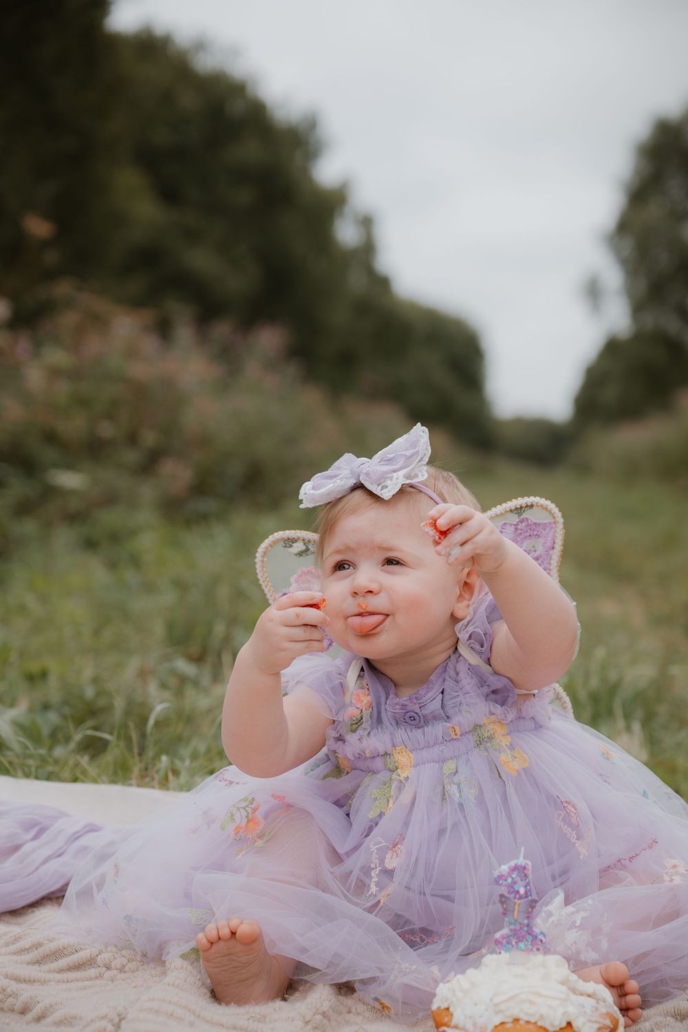 Baby girl in purple tutu dress with butterfly wings and bow headband eating cake outdoors on a blanket.
