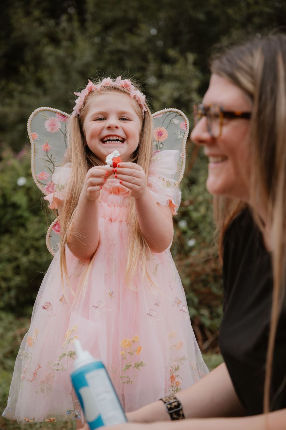 A joyful girl in a pink fairy costume with floral wings and headband laughs while holding a flower outdoors.