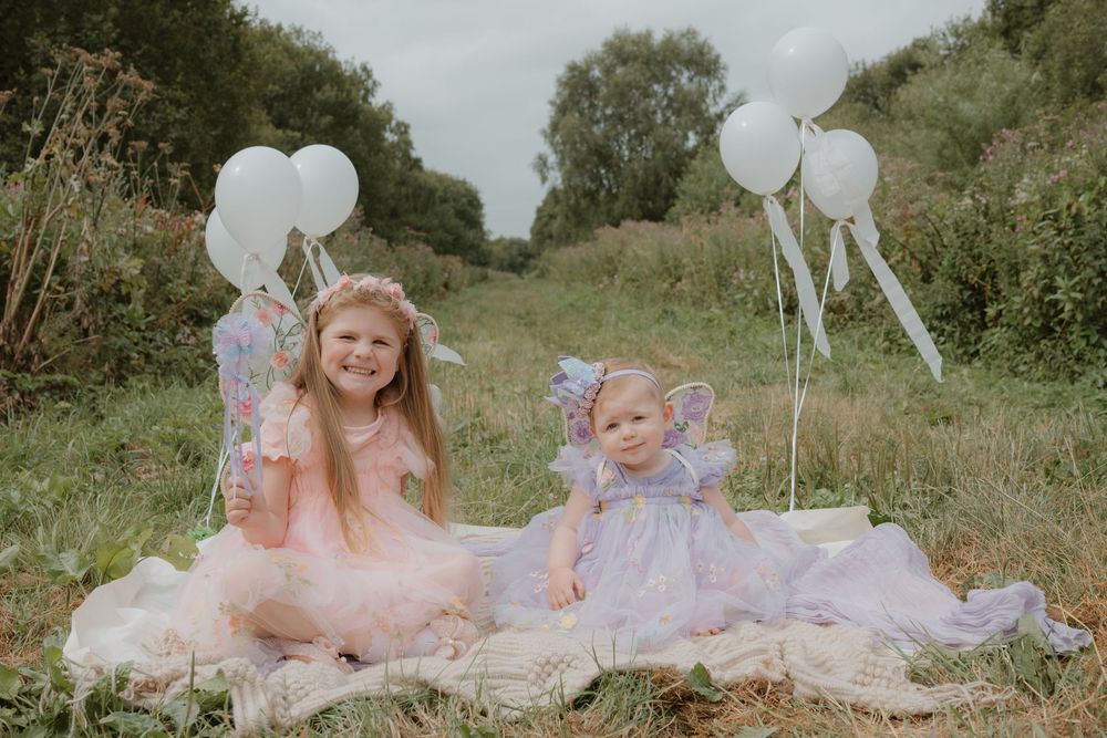 Two young girls in pastel fairy dresses sit on a blanket in a meadow, holding white balloons for a whimsical outdoor photoshoot.
