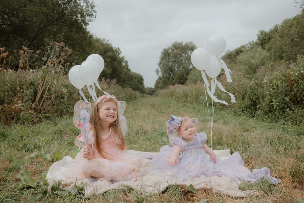 Two young girls in fairy costumes sit on a blanket in a meadow holding white heart-shaped balloons.