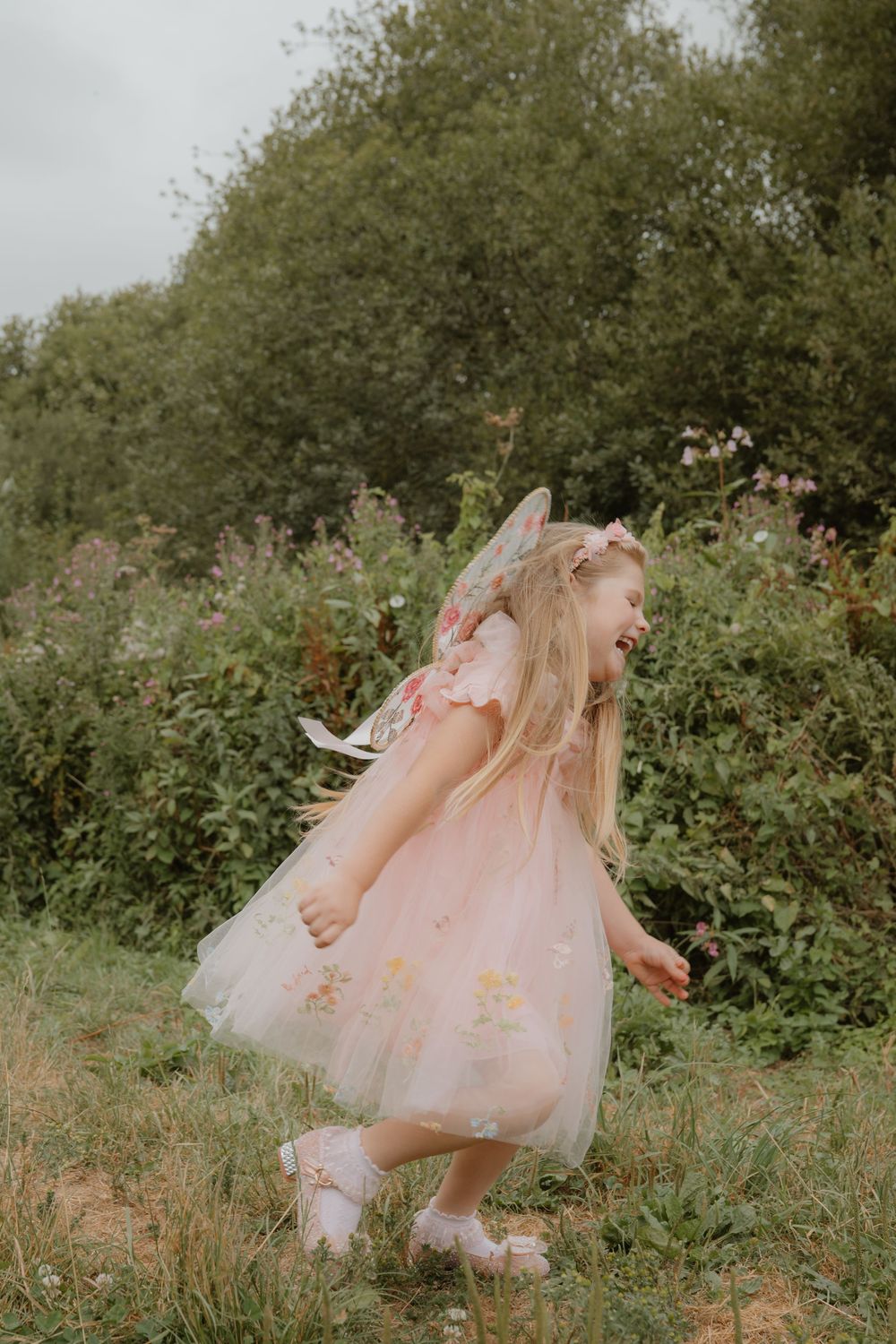 A joyful young girl in a pink tulle fairy dress with wings dances outdoors in a lush green garden.