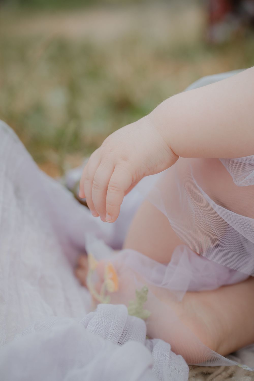 Close-up of a baby's tiny hand and chubby legs wrapped in a soft lavender tulle fabric outdoors.