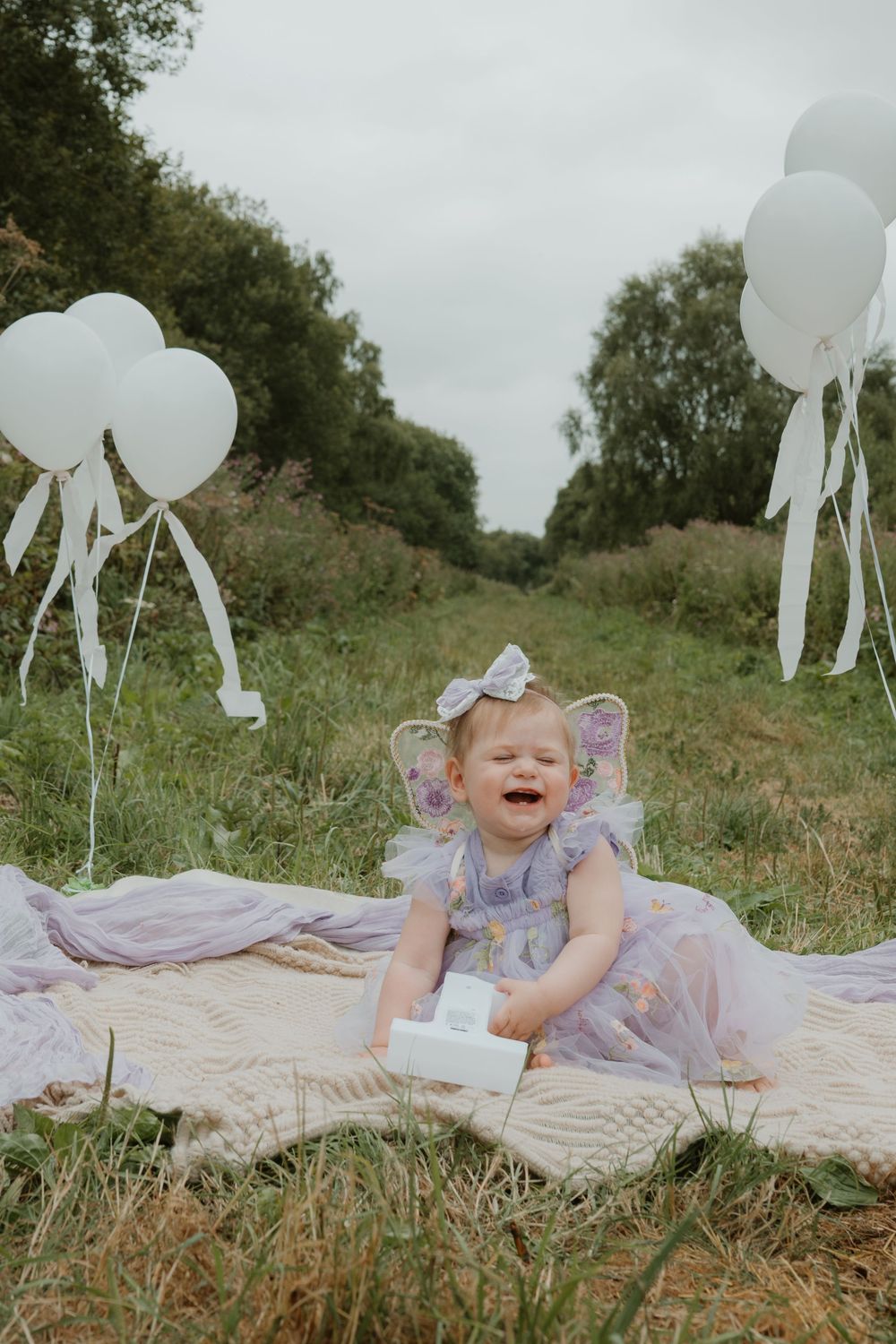 A crying baby girl in a purple fairy dress sits on a blanket outdoors surrounded by white balloons.