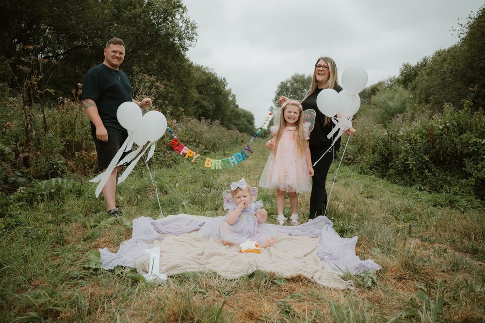 Family of four celebrating outdoors on a blanket with colorful banner and heart balloons in a green field.