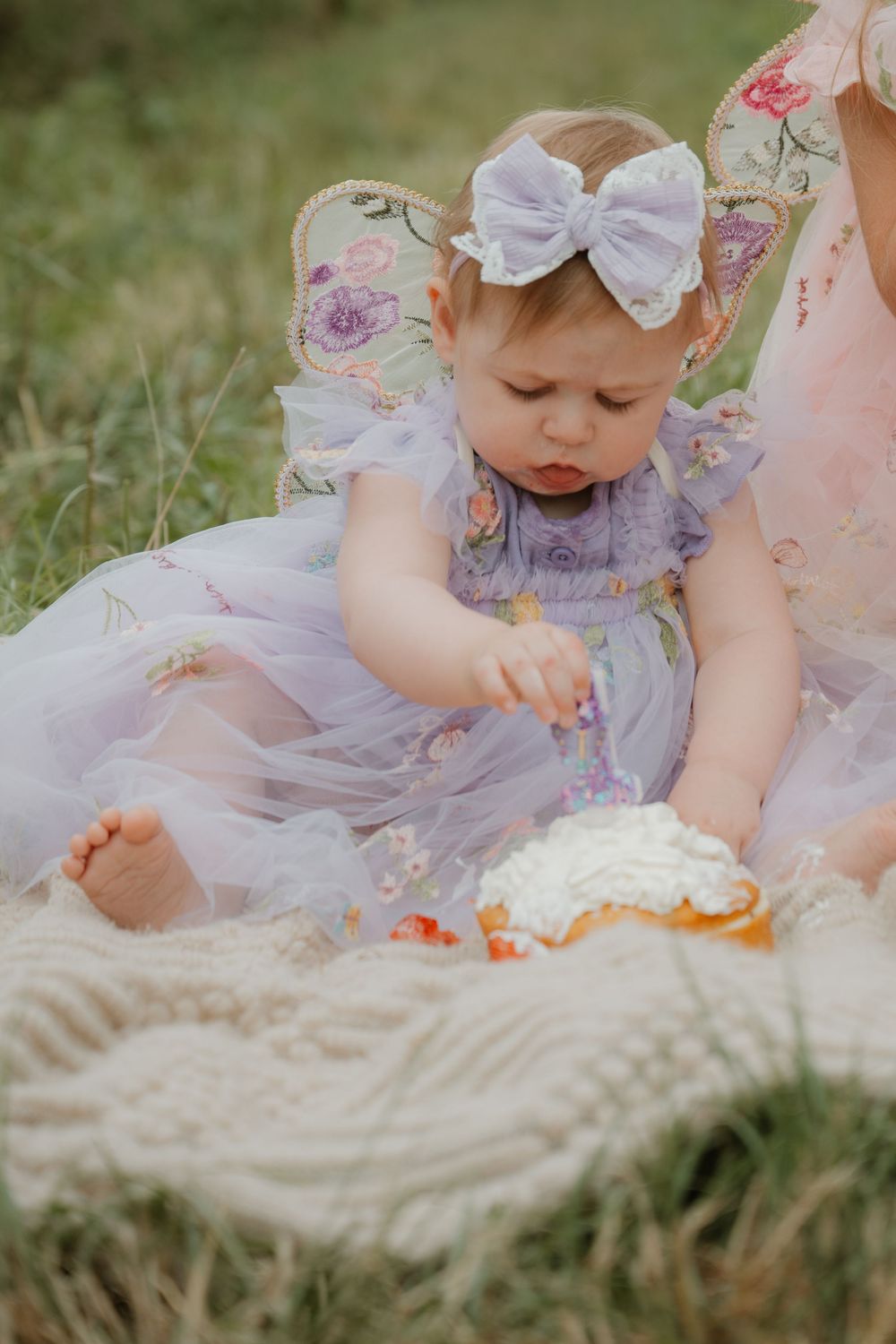 Baby girl in lavender tulle dress with bow headband smashing her first birthday cake outdoors on a blanket.