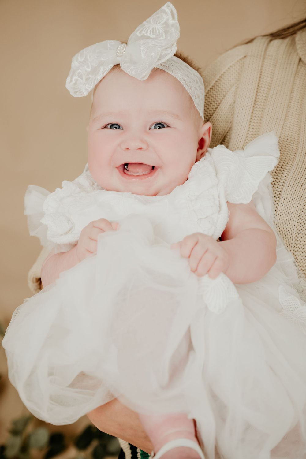 A baby in a white dress and bow headband smiling at the camera.