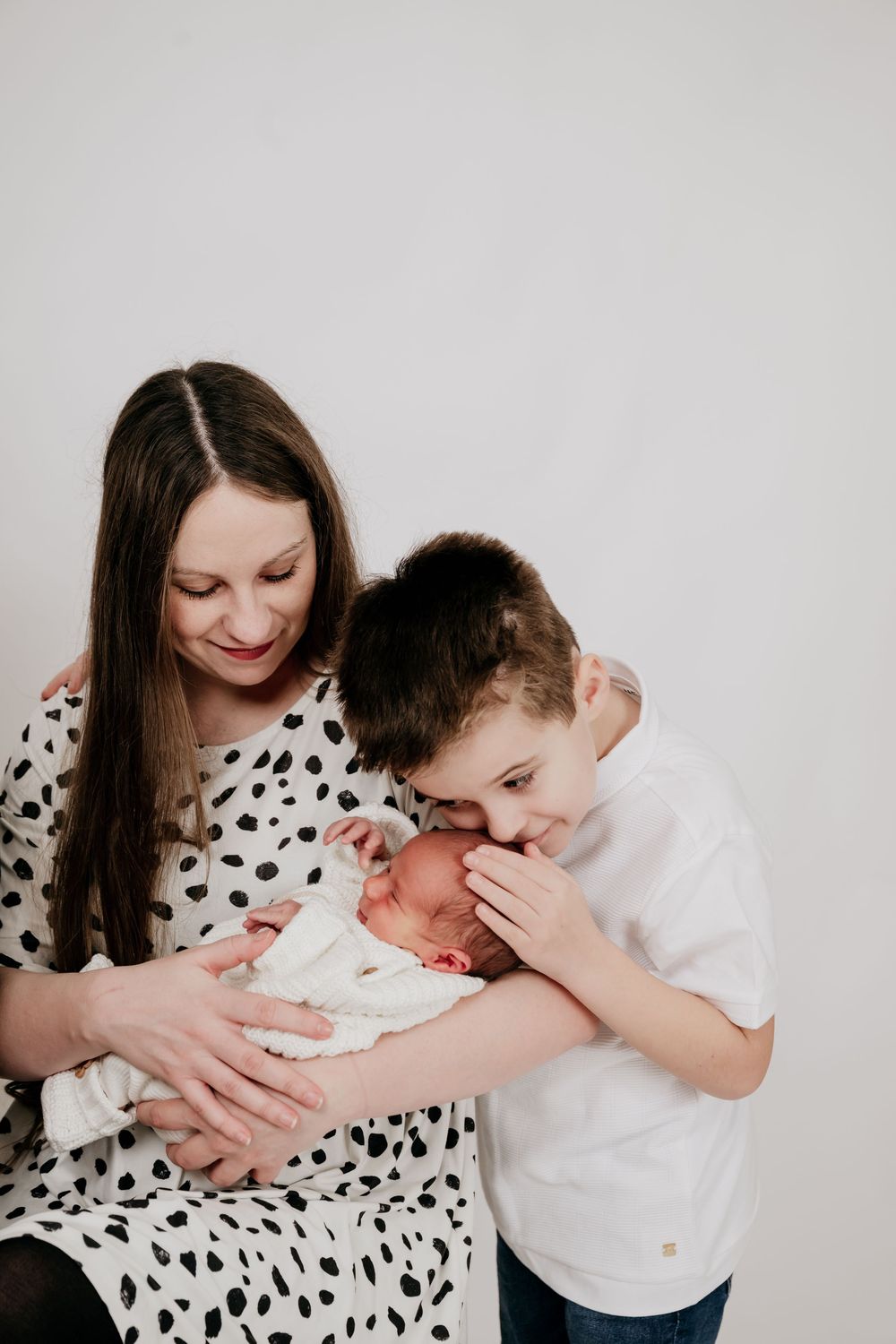 A mother in a polka dot dress holds her newborn baby while an older boy leans in to give the infant a gentle kiss.