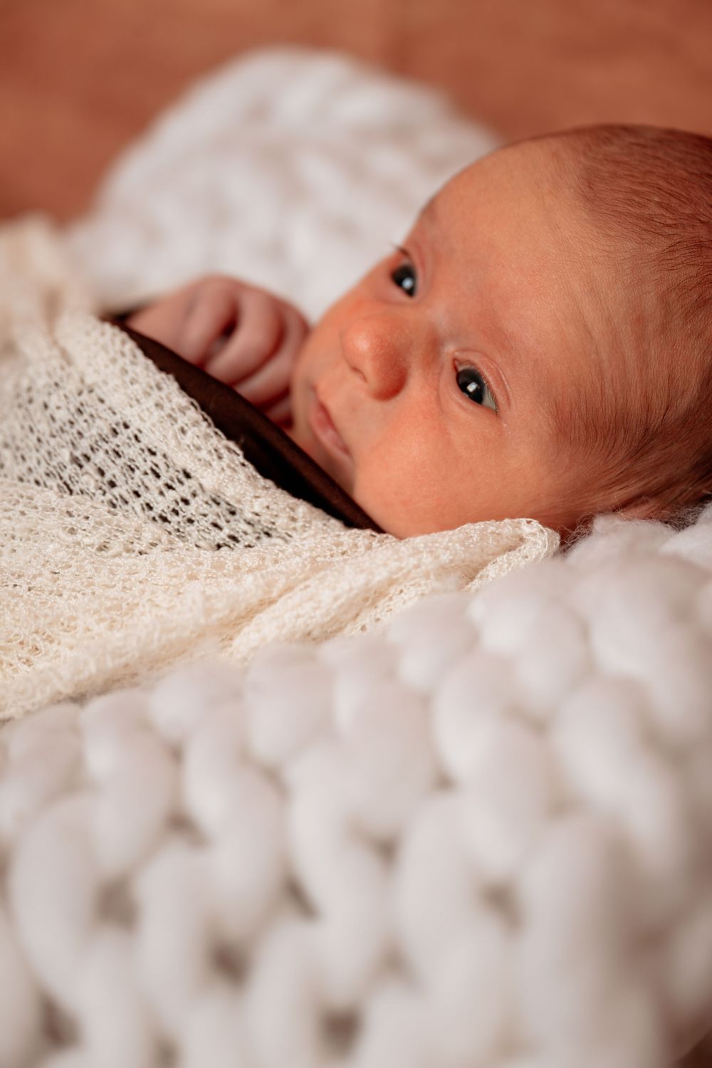 Newborn baby wrapped in a white knit blanket, lying on a chunky knit surface, with alert eyes open.