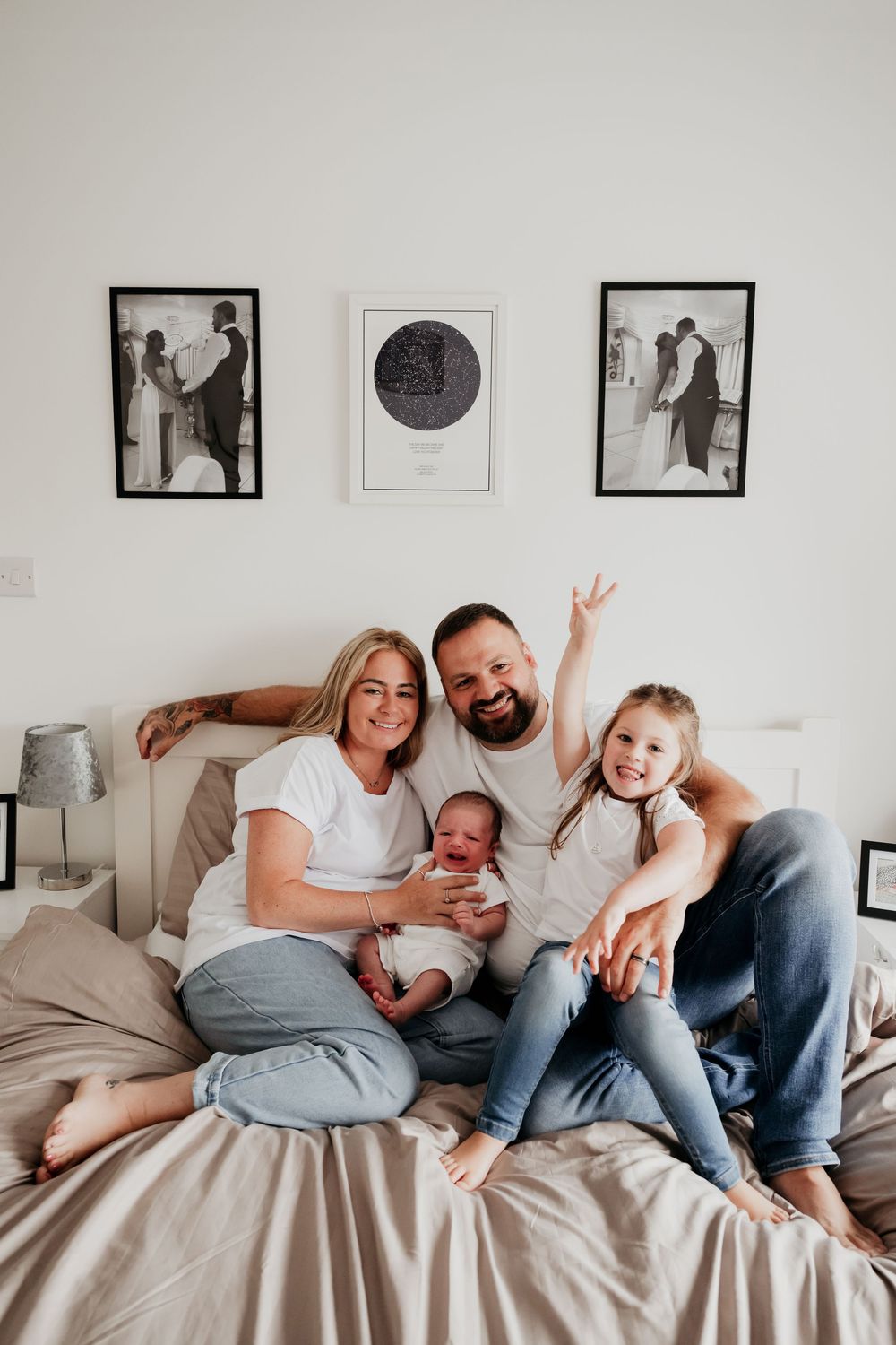Happy family of four sitting on bed, parents in white tees with newborn baby and excited toddler giving peace sign.