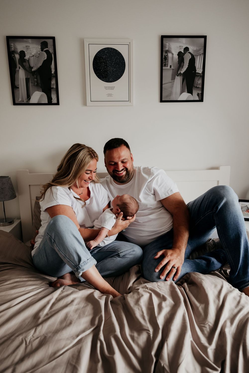 Happy couple sitting on bed laughing with newborn baby, framed photos on wall behind them.