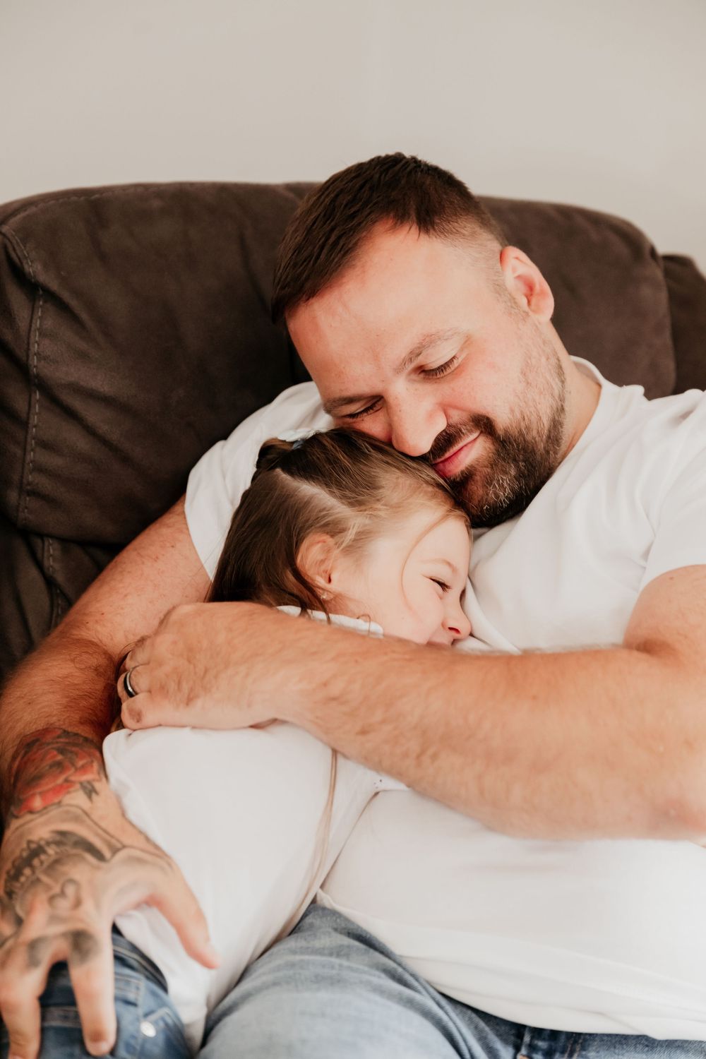A smiling father with tattoos hugs his young daughter on a brown sofa.