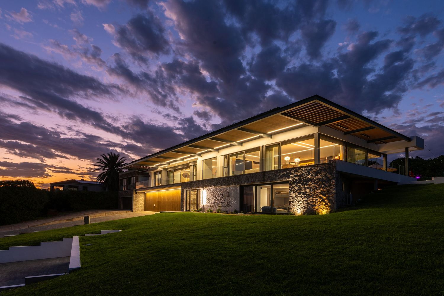 Contemporary glass house illuminated at twilight with stunning colorful cloudy sunset backdrop.
