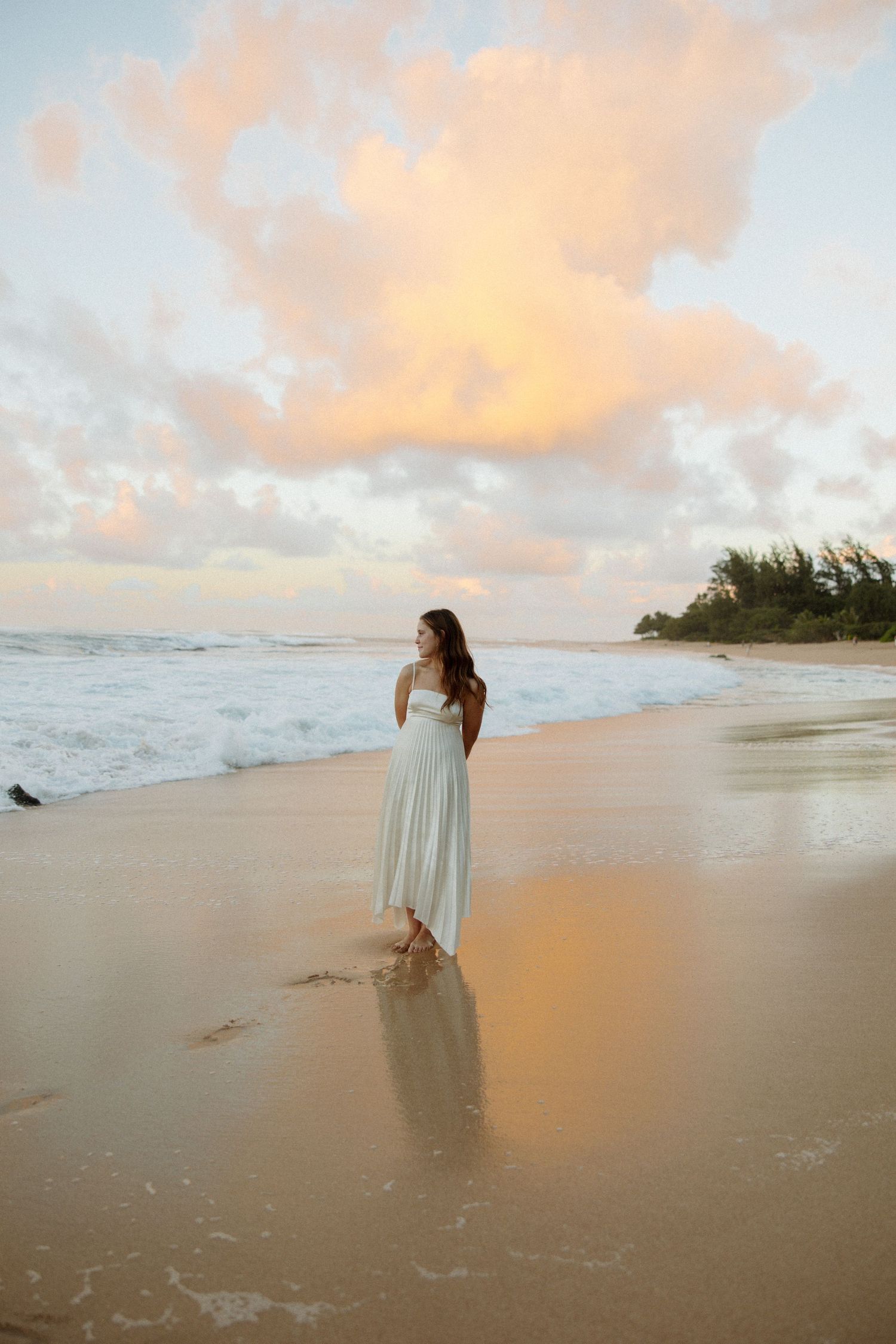A woman in a light dress stands on a beach at sunset with waves and palm trees in the distance.
