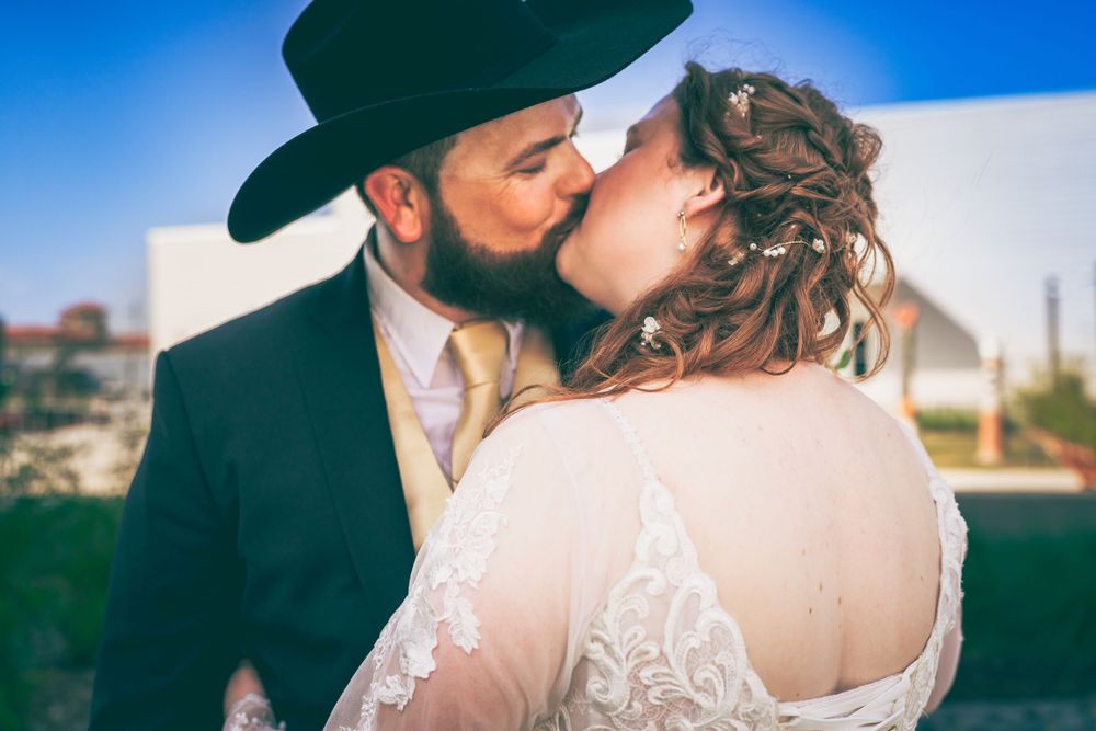 A tender moment between newlyweds captured in beautiful afternoon light, with her lace gown and his black hat silhouetted against blue sky.