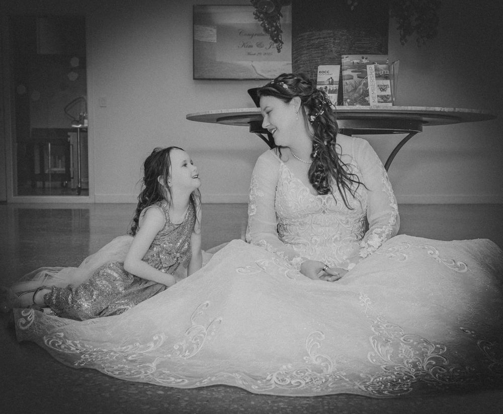 A child and adult wearing formal dresses sit together sharing a playful moment in black and white photography.