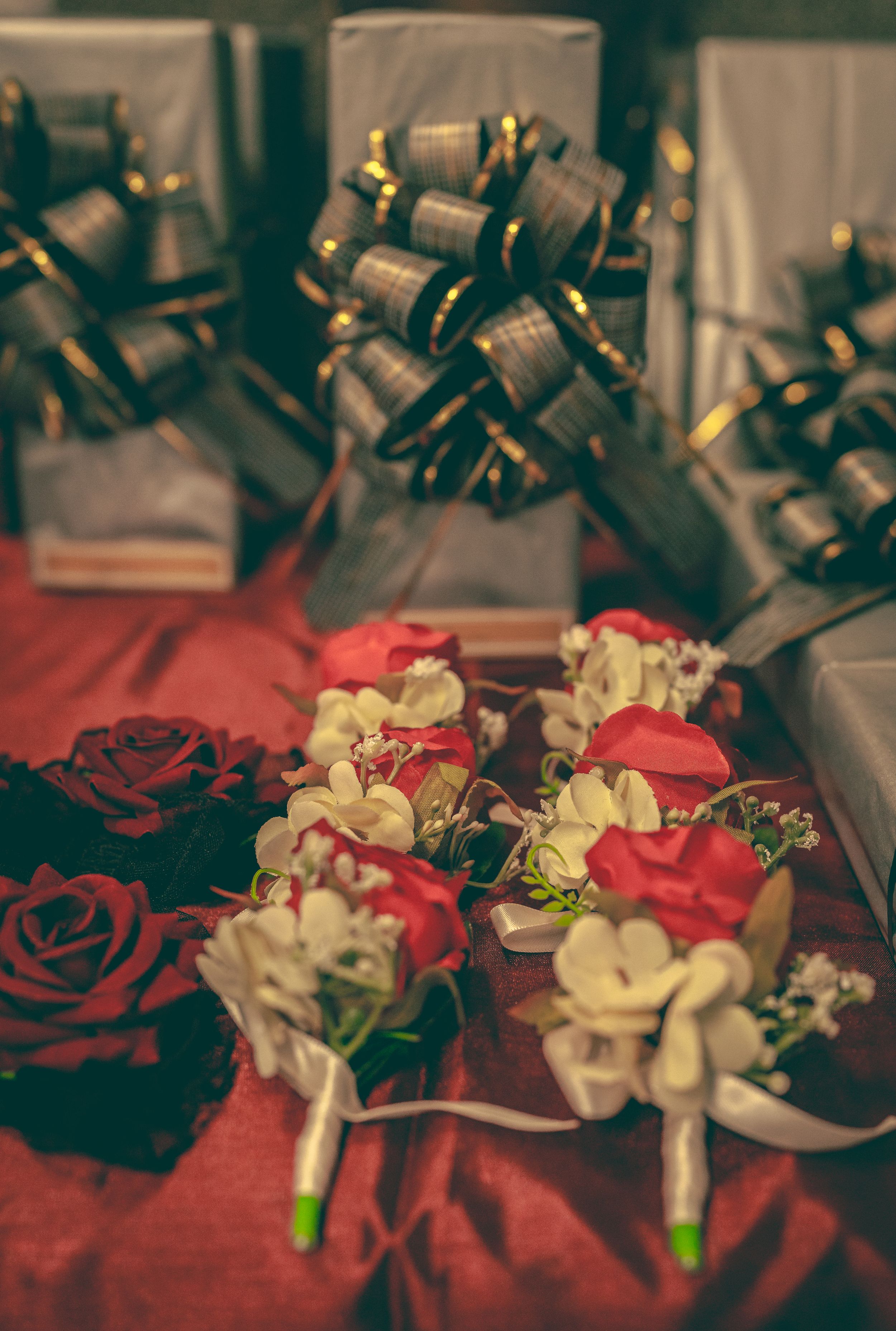 Red and white wedding boutonnieres with roses and ribbons displayed beside wrapped gifts on a red surface.