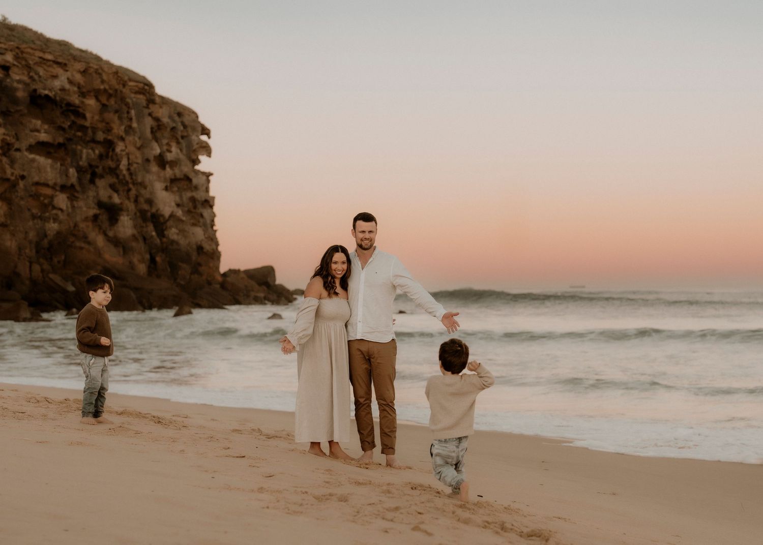 Family of four on beach at pink sunset, parents holding hands with two young boys.