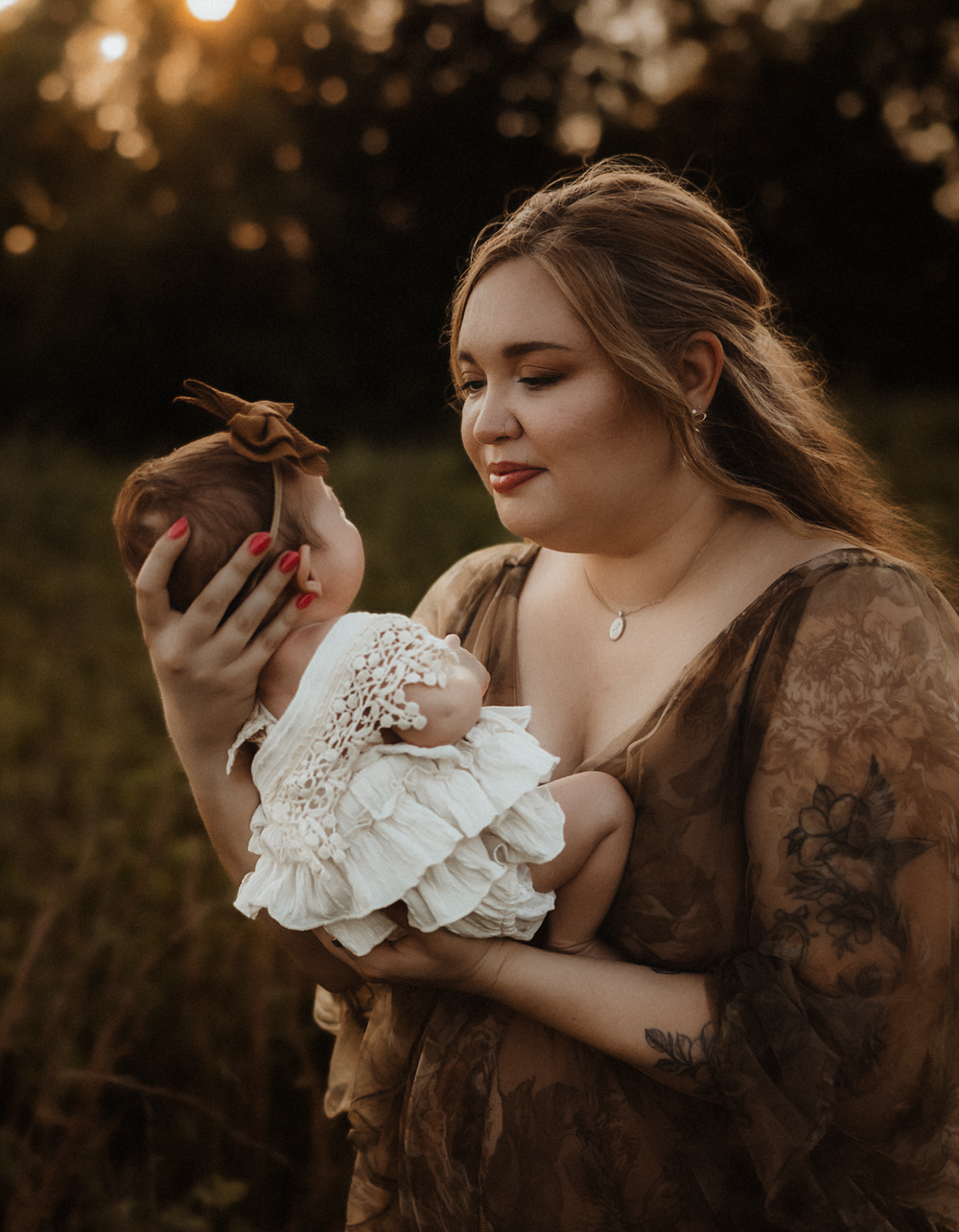 A young mother in a brown lace dress holds her newborn baby in white lace at golden hour outdoors.