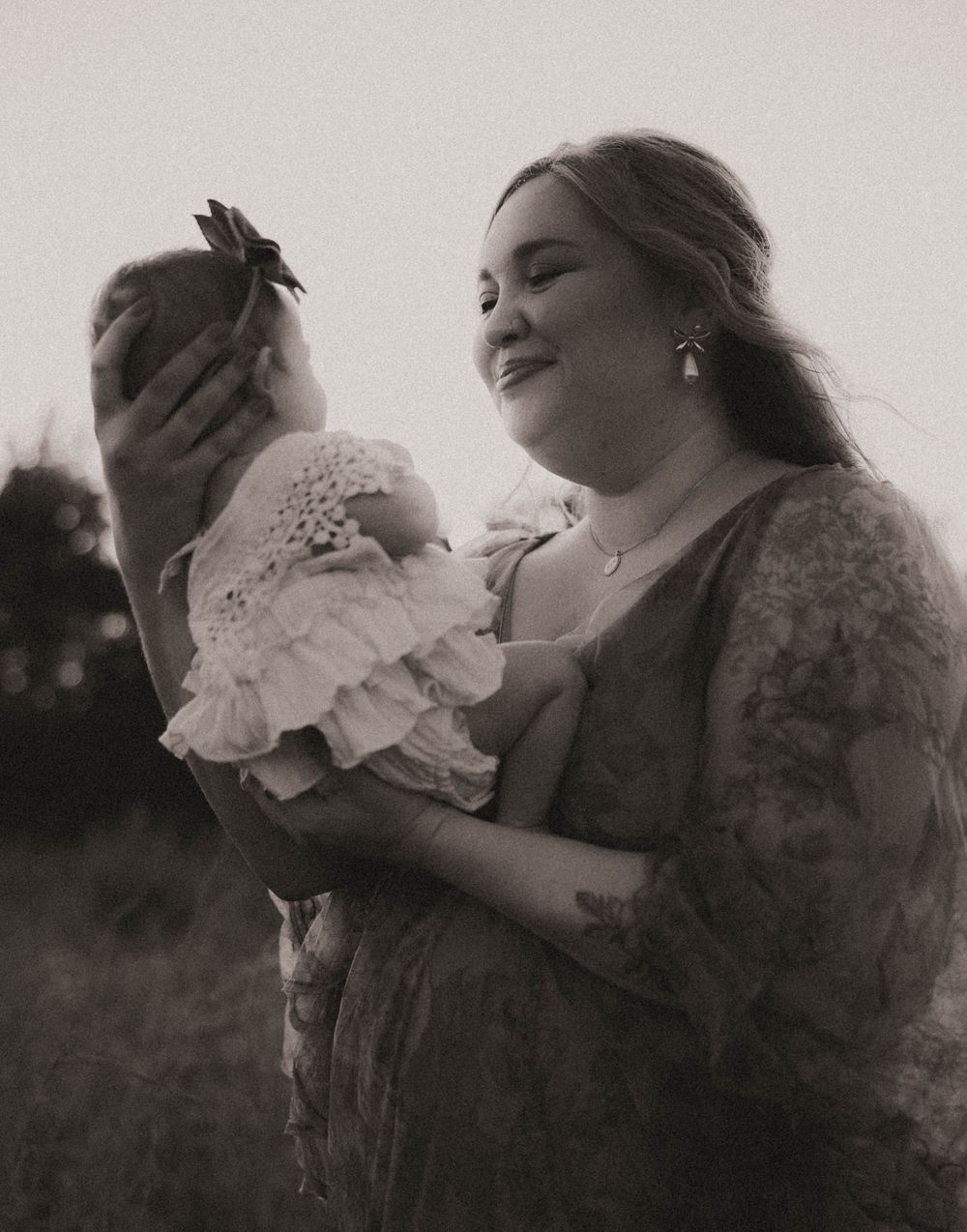 Black and white photo of a smiling mother holding her newborn baby dressed in a white lace outfit outdoors.