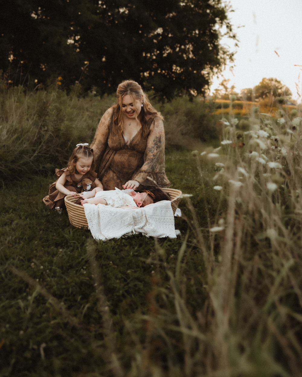A mother and young daughter admire a newborn baby in a wicker basket in a lush green meadow at golden hour.