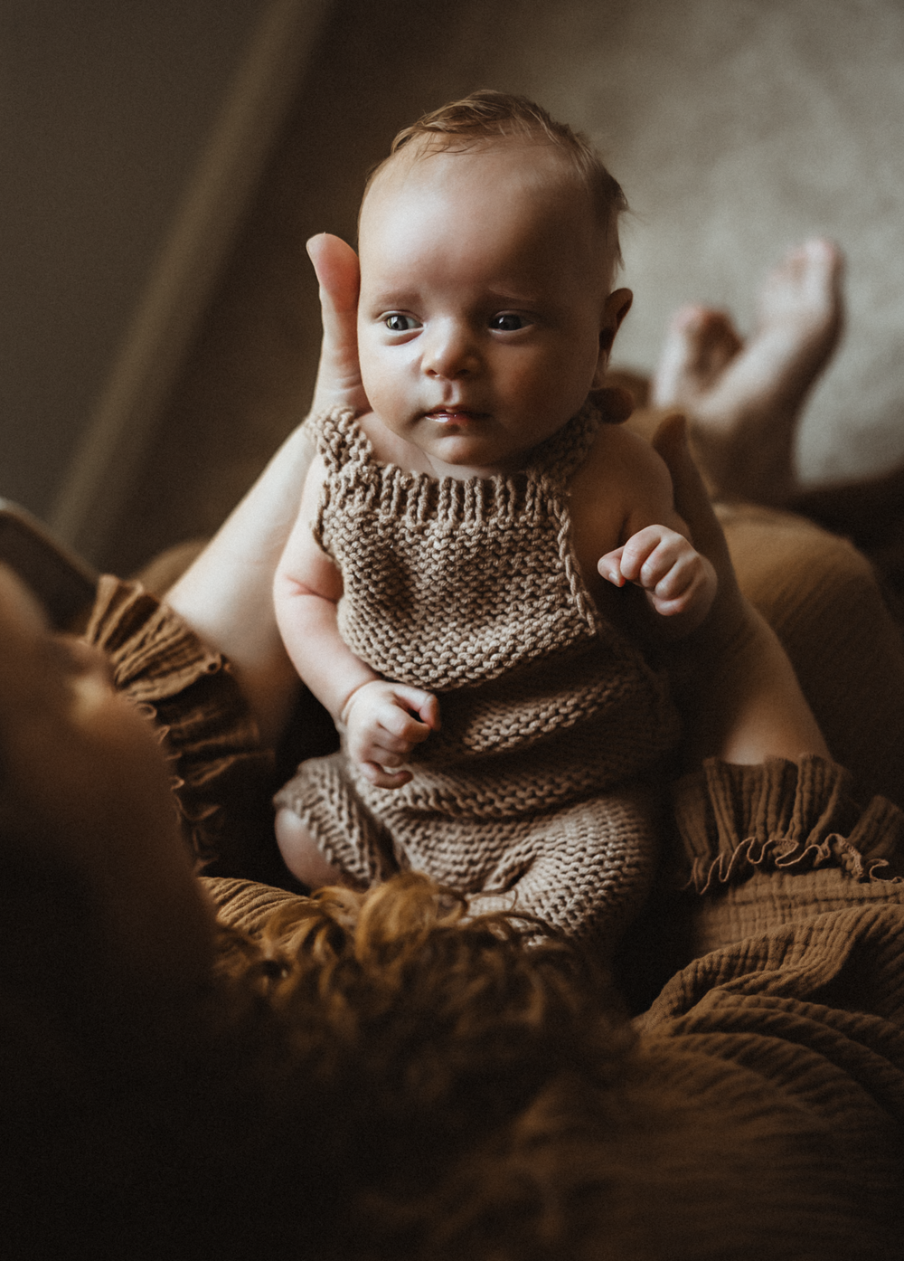 A newborn baby in a knit romper being held upright by a parent against warm golden fabric.
