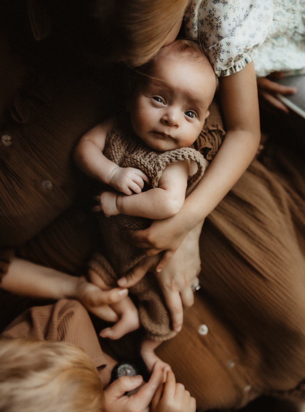 A newborn baby in a knit romper being cradled by a mother while an older sibling reaches up to touch the infant.