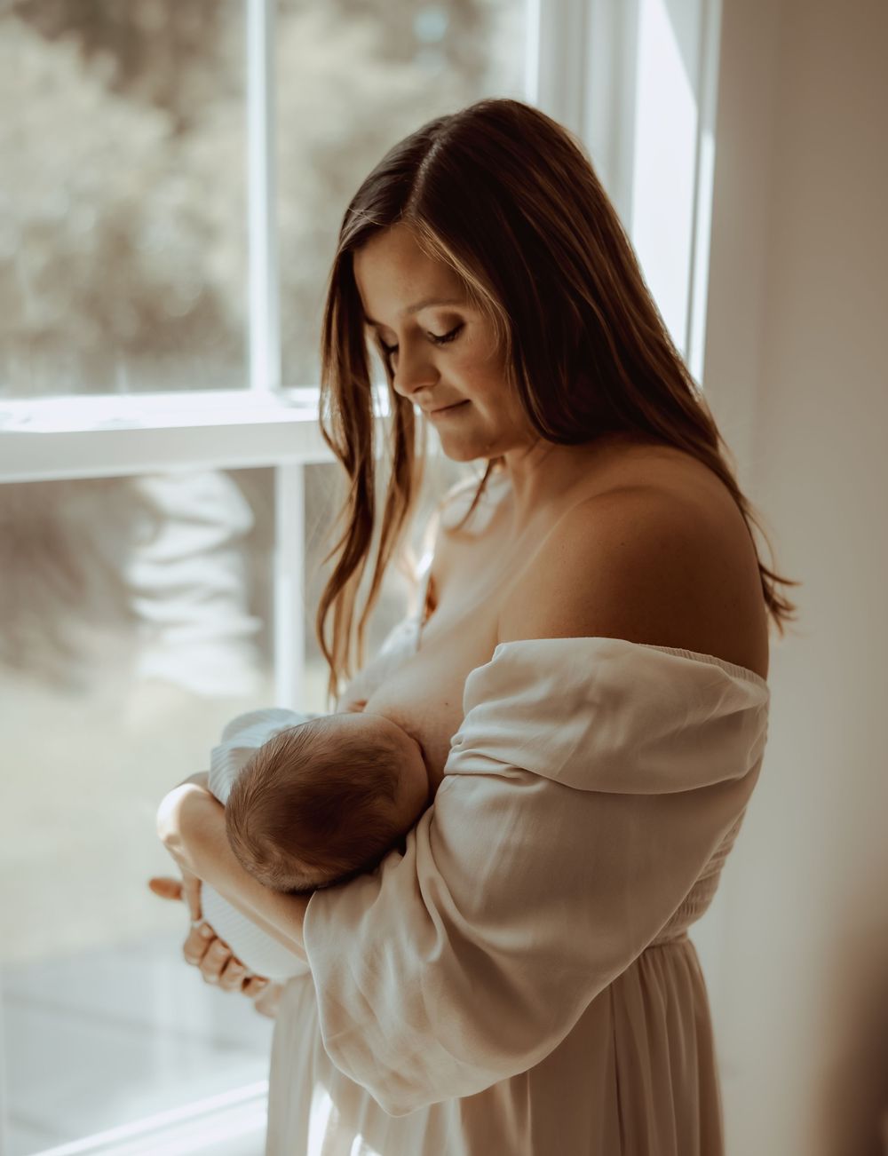 A mother tenderly breastfeeds her newborn baby by a sunlit window, wearing an off-shoulder white dress.