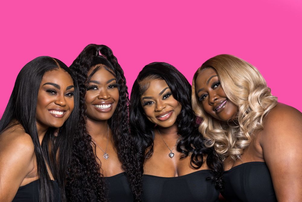 Four friends in black dresses pose together against a vibrant pink background for a glamorous portrait.