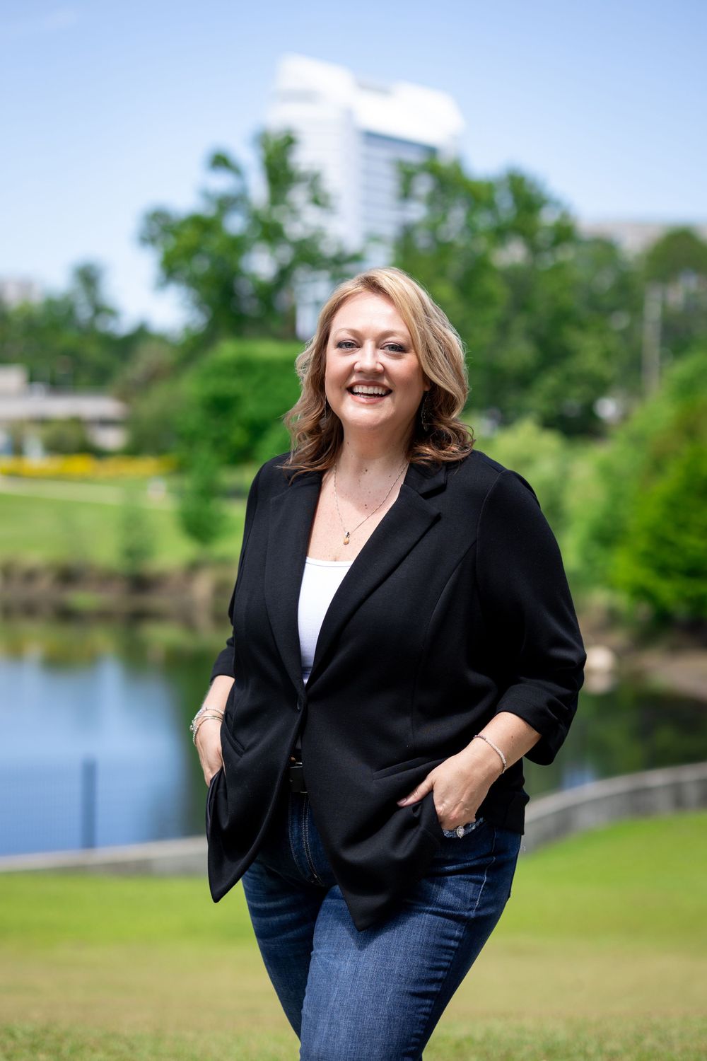 A woman in a black blazer smiles outdoors with a lake and trees in the background.