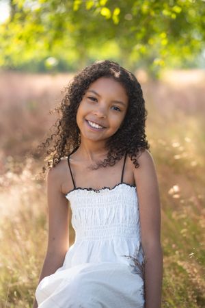 A portrait of a person in a white dress sitting in a sunlit field with curly hair.