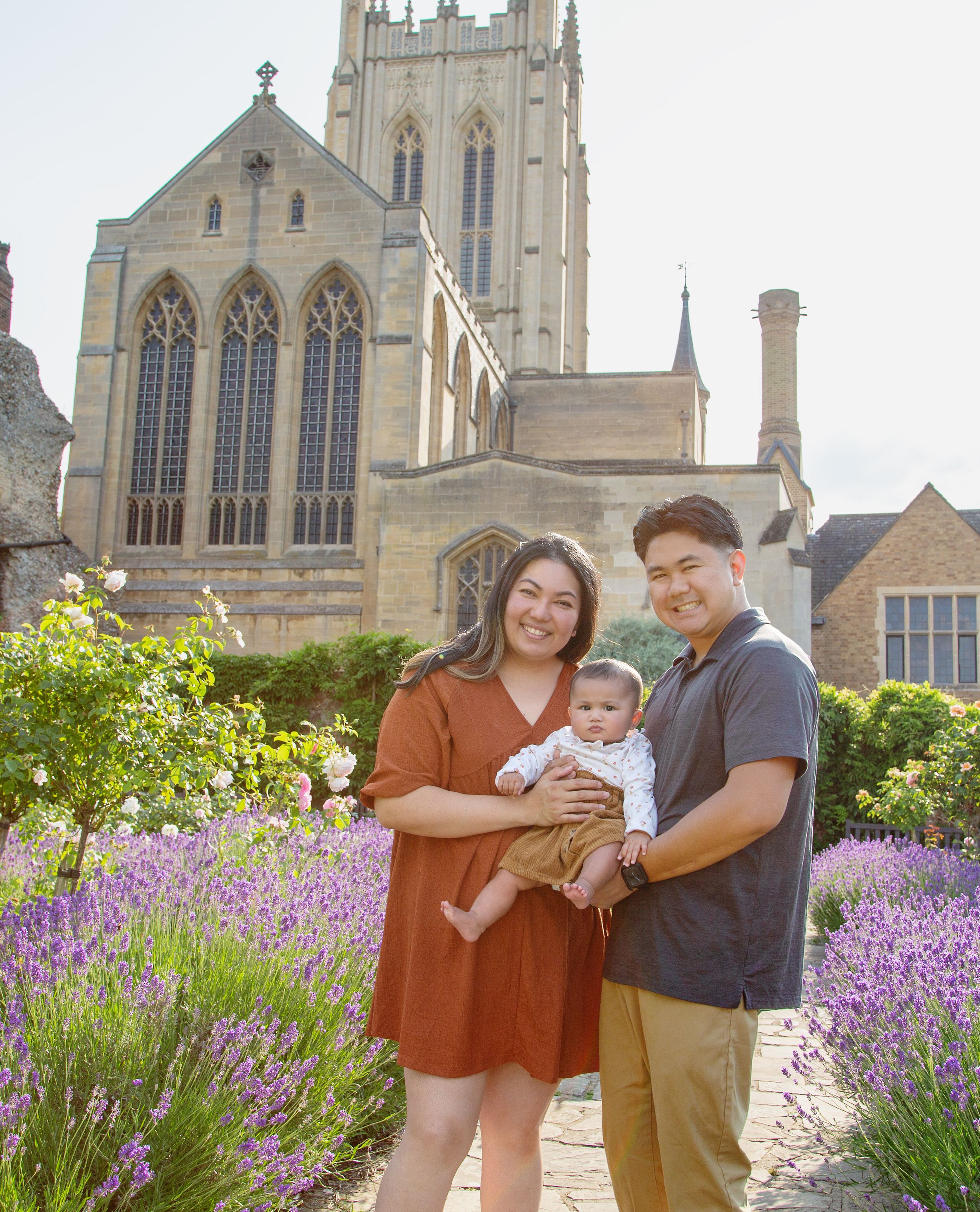 A young family poses together in front of a Gothic church with blooming lavender gardens.