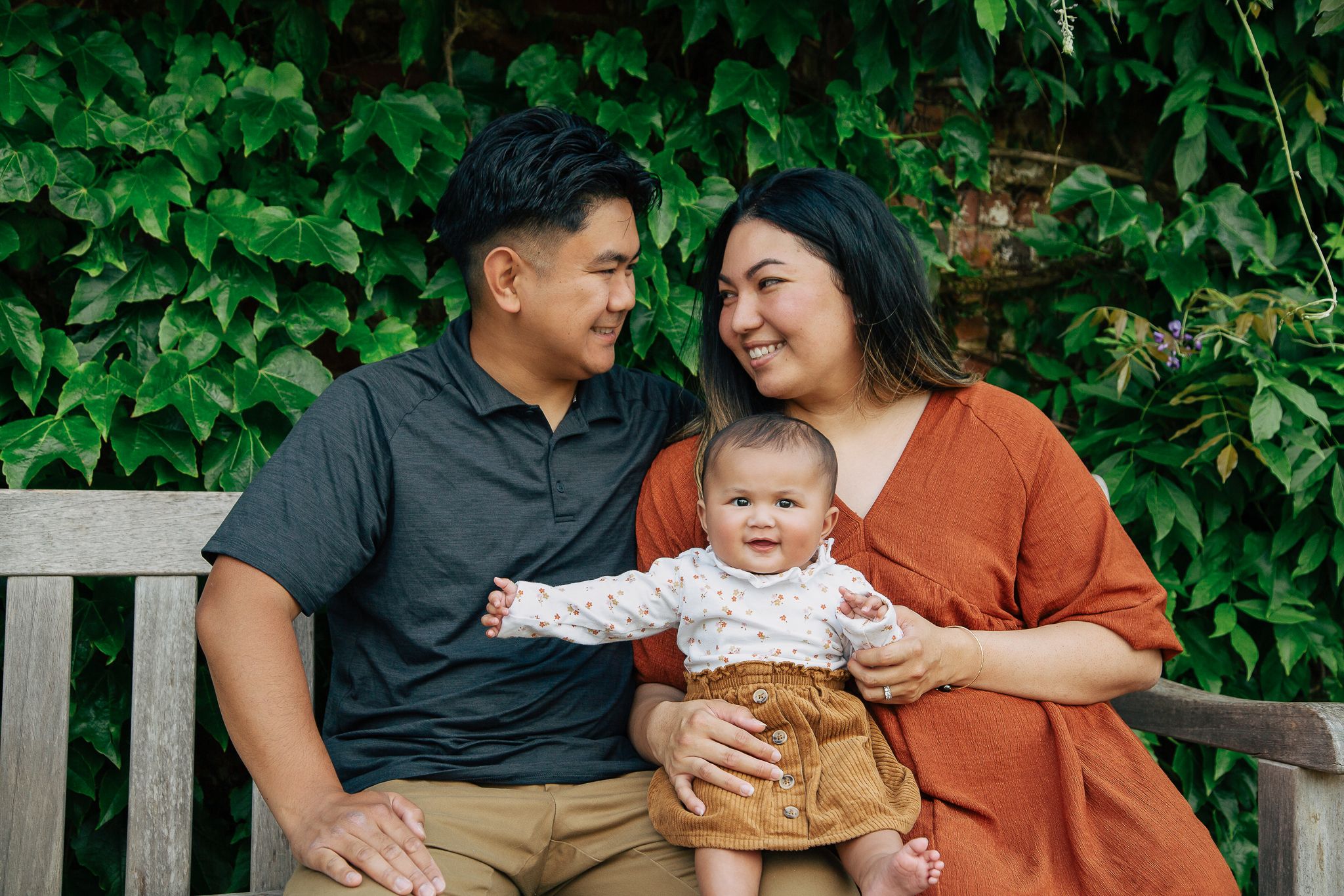 A family sits together on a wooden bench against an ivy-covered wall.