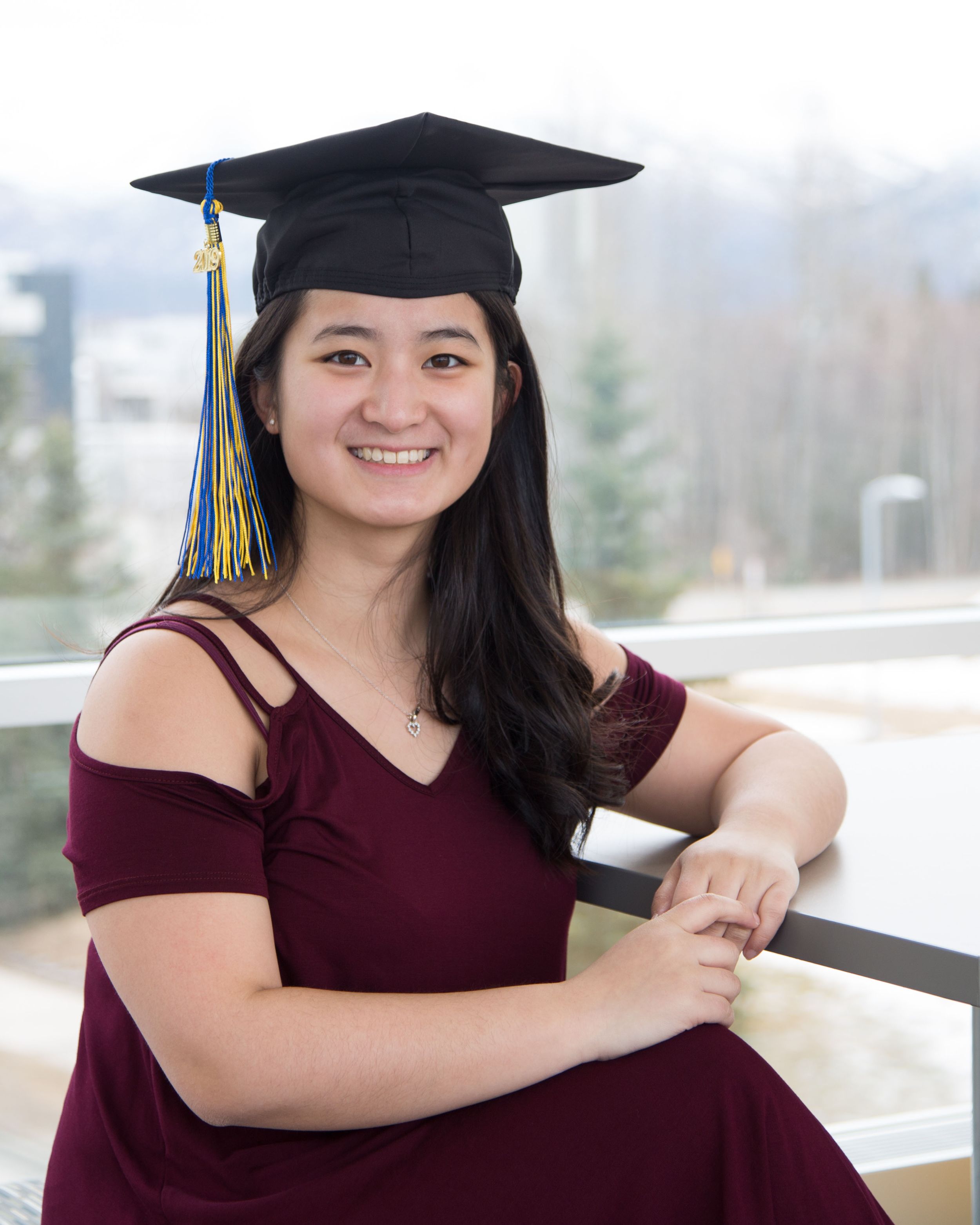 Graduate in burgundy dress poses with cap and tassel during outdoor graduation photo session.