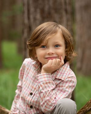 Portrait of a child in a checkered shirt resting against a tree in a pine forest.