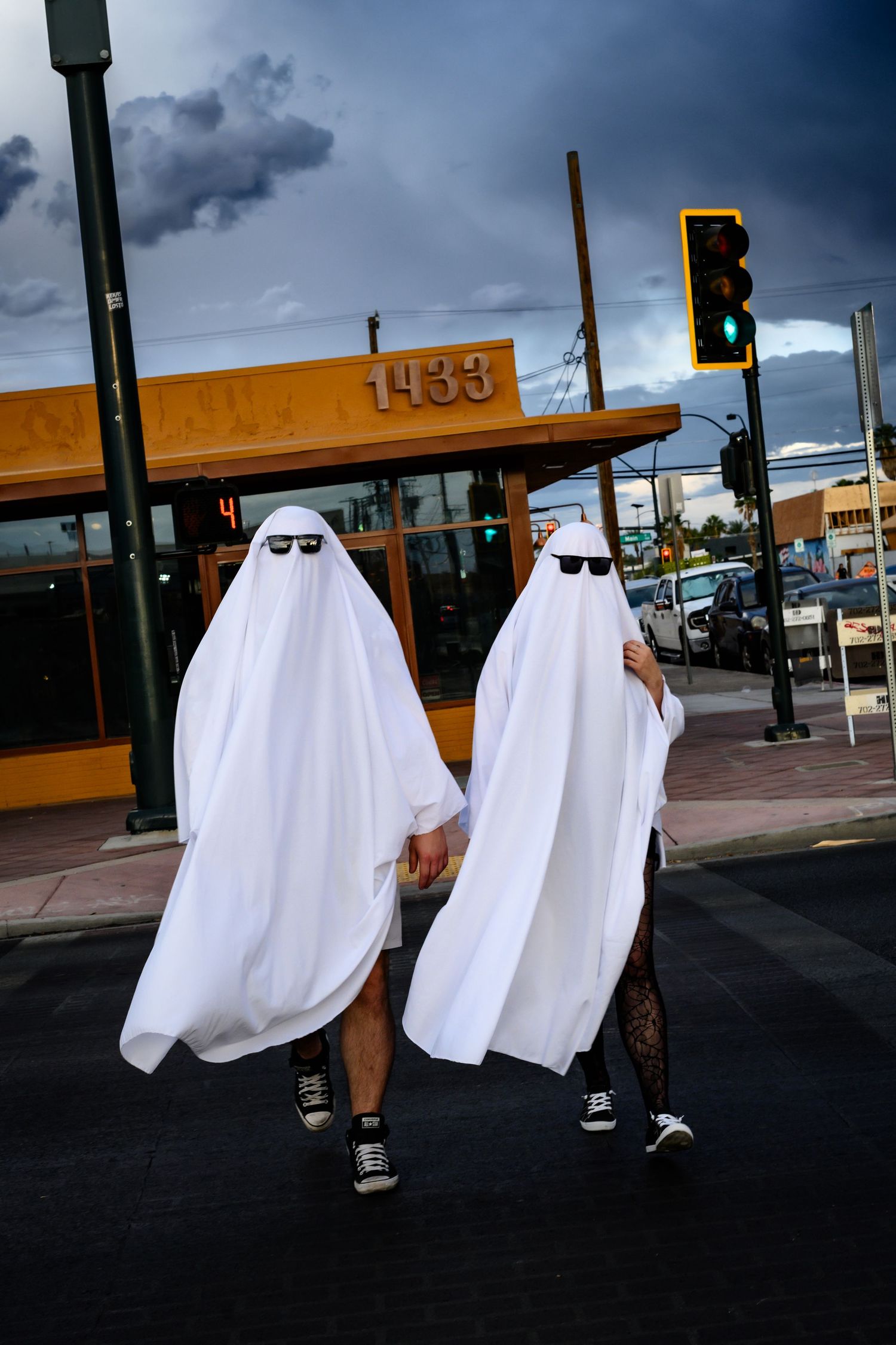 Two figures in ghost costumes walking past an orange building under a dramatic cloudy sky.