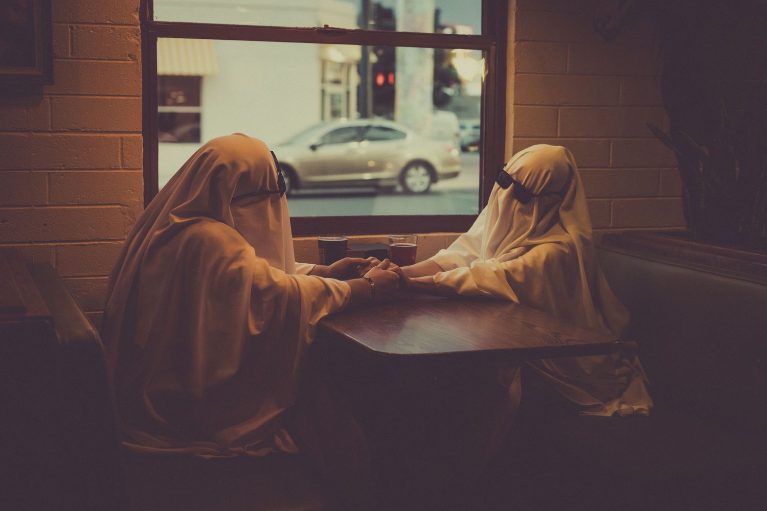 Two figures in golden hooded robes sit across from each other at a window table in a dimly lit room.